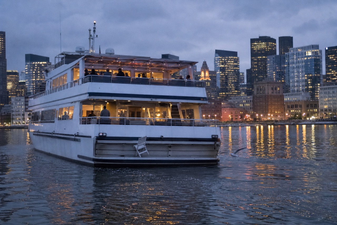 A dinner cruise vessel underway on Boston Harbor at dusk, city lights reflected on the water