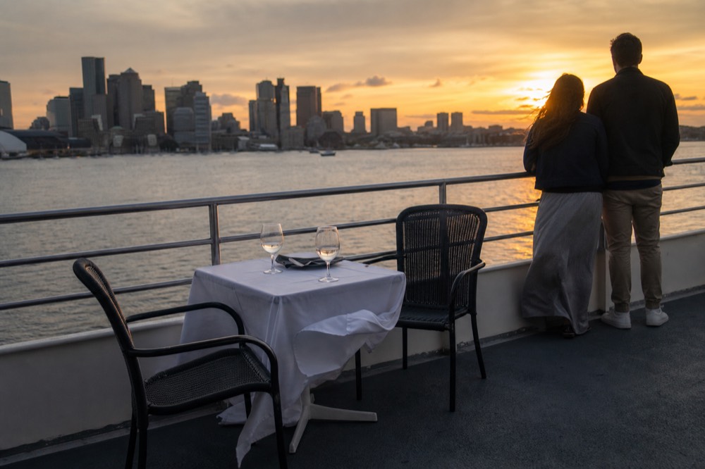 A couple at a dinner table on a cruise deck with the Boston skyline behind them