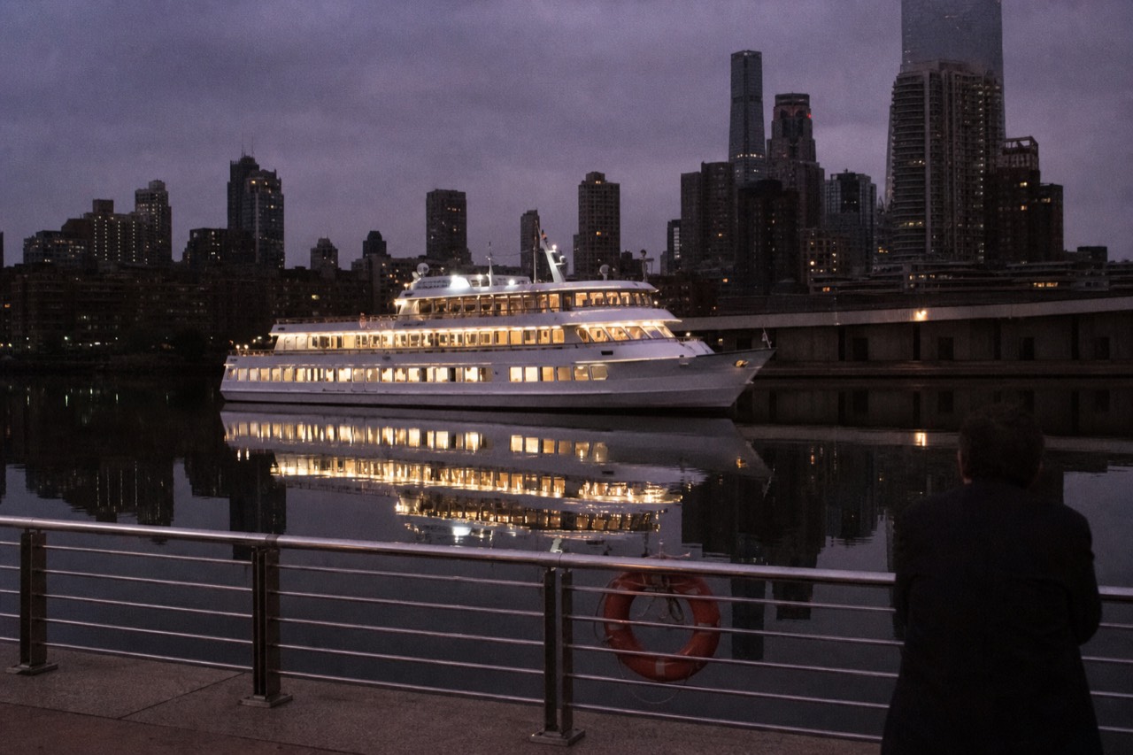 A lit dinner cruise vessel on Boston Harbor at dusk with the city skyline behind it