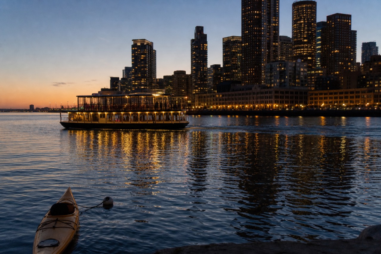 A dinner cruise vessel lit up at dusk on Boston Harbor, with the city skyline behind it