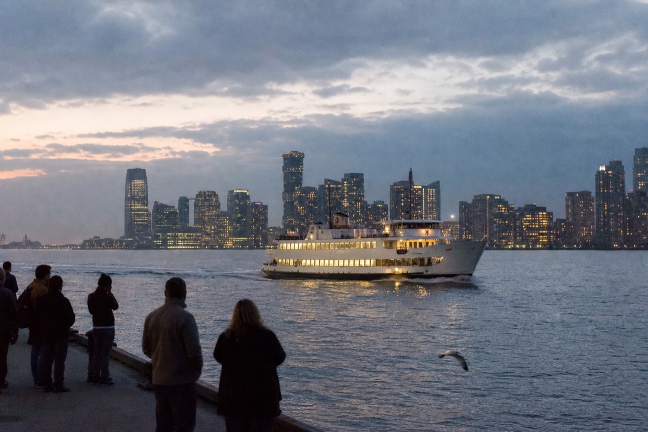 An illuminated dinner cruise vessel on Boston Harbor at dusk with the downtown skyline behind it