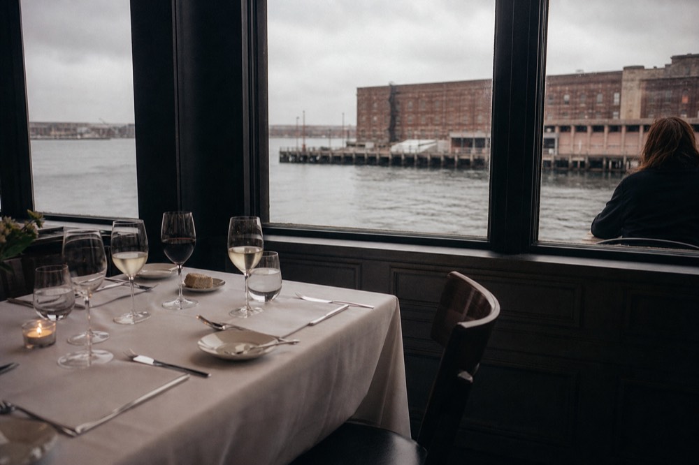 A set dinner table with glassware beside a window overlooking the harbor