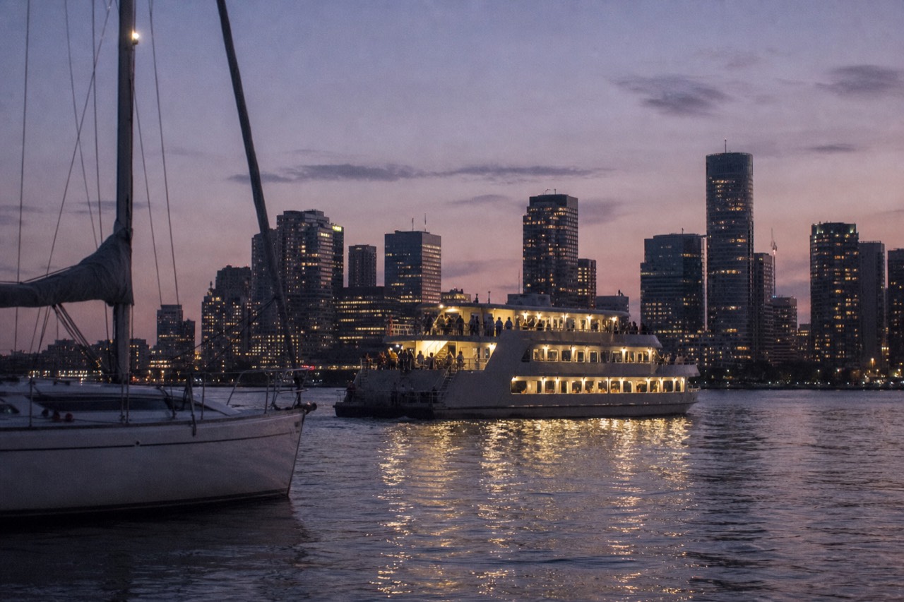 A dinner cruise vessel on Boston Harbor at dusk with the city skyline behind it