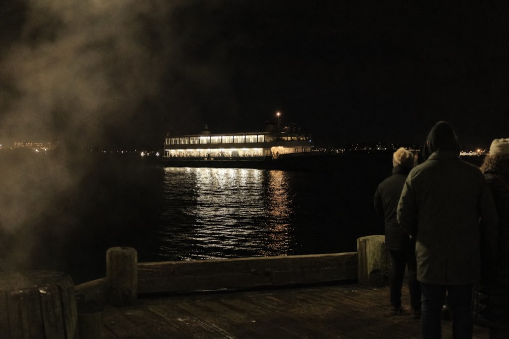 A harbor dinner cruise vessel lit at night seen from a Boston pier