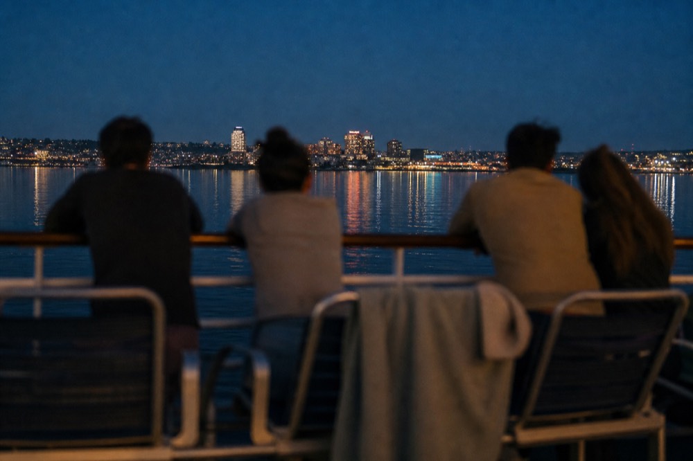 Passengers on an exterior deck of a dinner cruise watching the Boston skyline light up after sunset