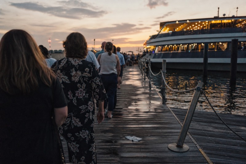 Passengers boarding a lit buffet dinner cruise boat at a Boston pier on a summer evening