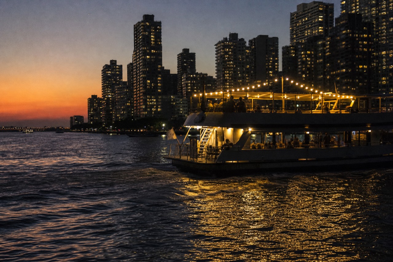 Dinner cruise vessel lit up at dusk on Boston Harbor with the city skyline behind