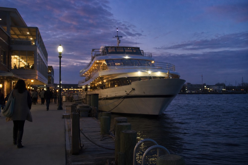A dinner cruise vessel lit at dusk on Boston Harbor with the city skyline behind it