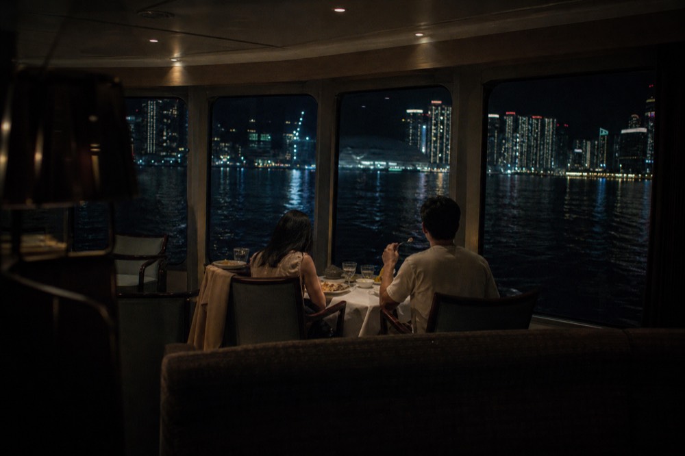 A couple sharing a plated dinner at a window table on a cruise ship, city lights on the water beyond