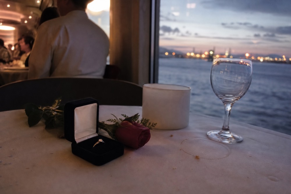 An engagement ring box beside a rose on a dinner cruise table with the harbor in the background