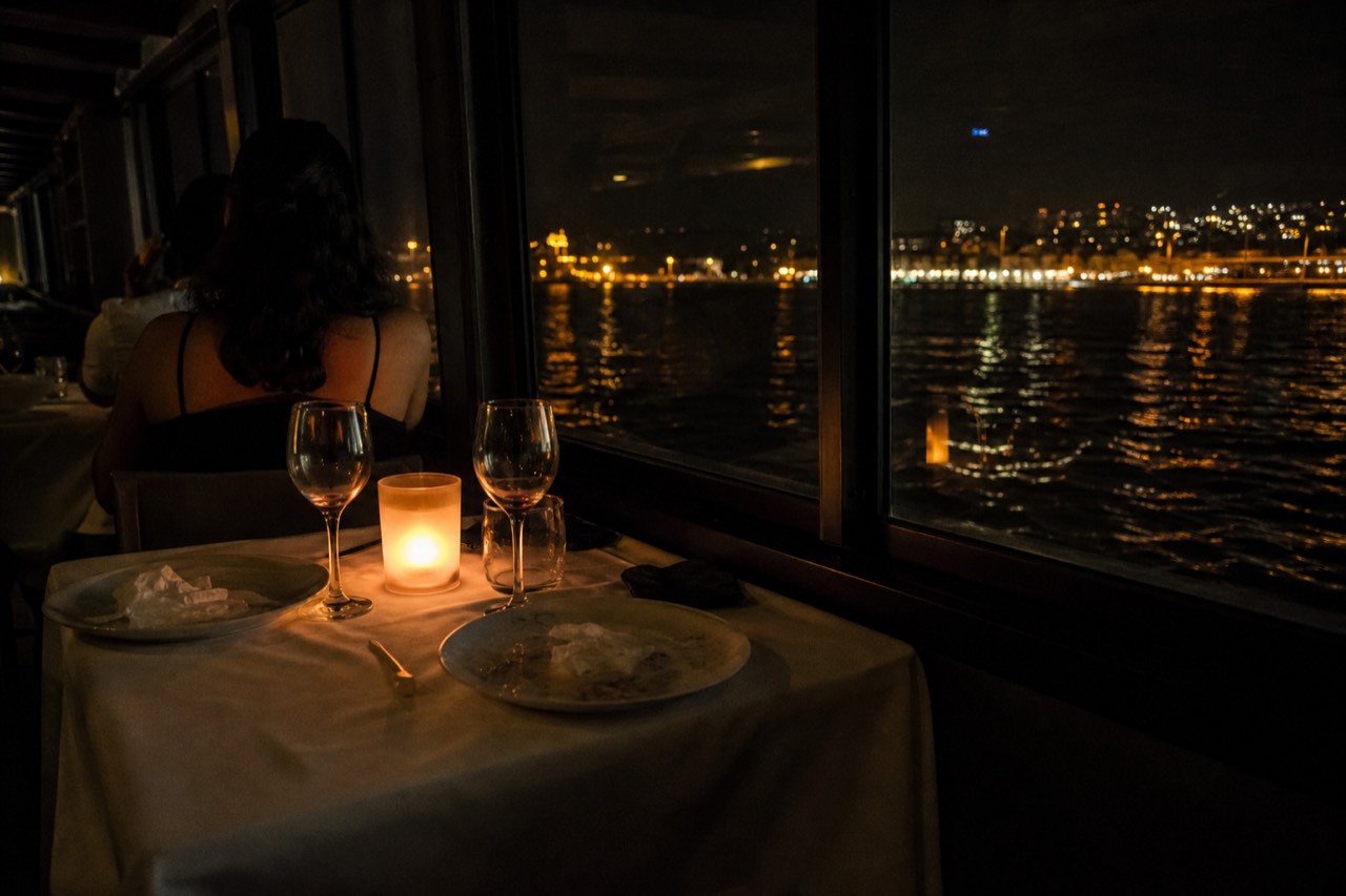 A couple at a candlelit window table on an evening harbor cruise with city lights on the water beyond
